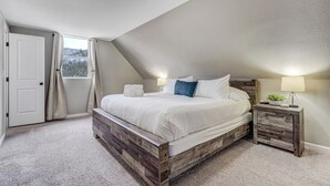Une chambre avec un plafond en pente, un cadre de lit en bois, une table de chevet avec une lampe, et une vue sur une montagne par la fenêtre.