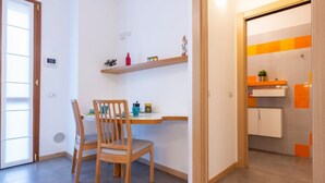A room with a wooden chair, a small table, a shelf with items, and a door leading to a bathroom with orange tiles. - GuestHost - Just a few minutes’ walk from the subway, in the heart of the town’s historic center, Casa Klimt is a quiet, renovated two-room apartment in an old house, inspired by the colors and landscapes of the famous painter. Filled with natural li (Cernusco sul Naviglio)
