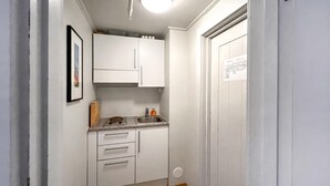 A small, narrow kitchen with white cabinets, a countertop, and a sink. There is a framed picture on the wall and a door with a sign on it.
