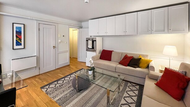 A modern living room with a glass coffee table, a beige sofa with red and yellow cushions, a white wall with a framed picture, and a built-in oven.
