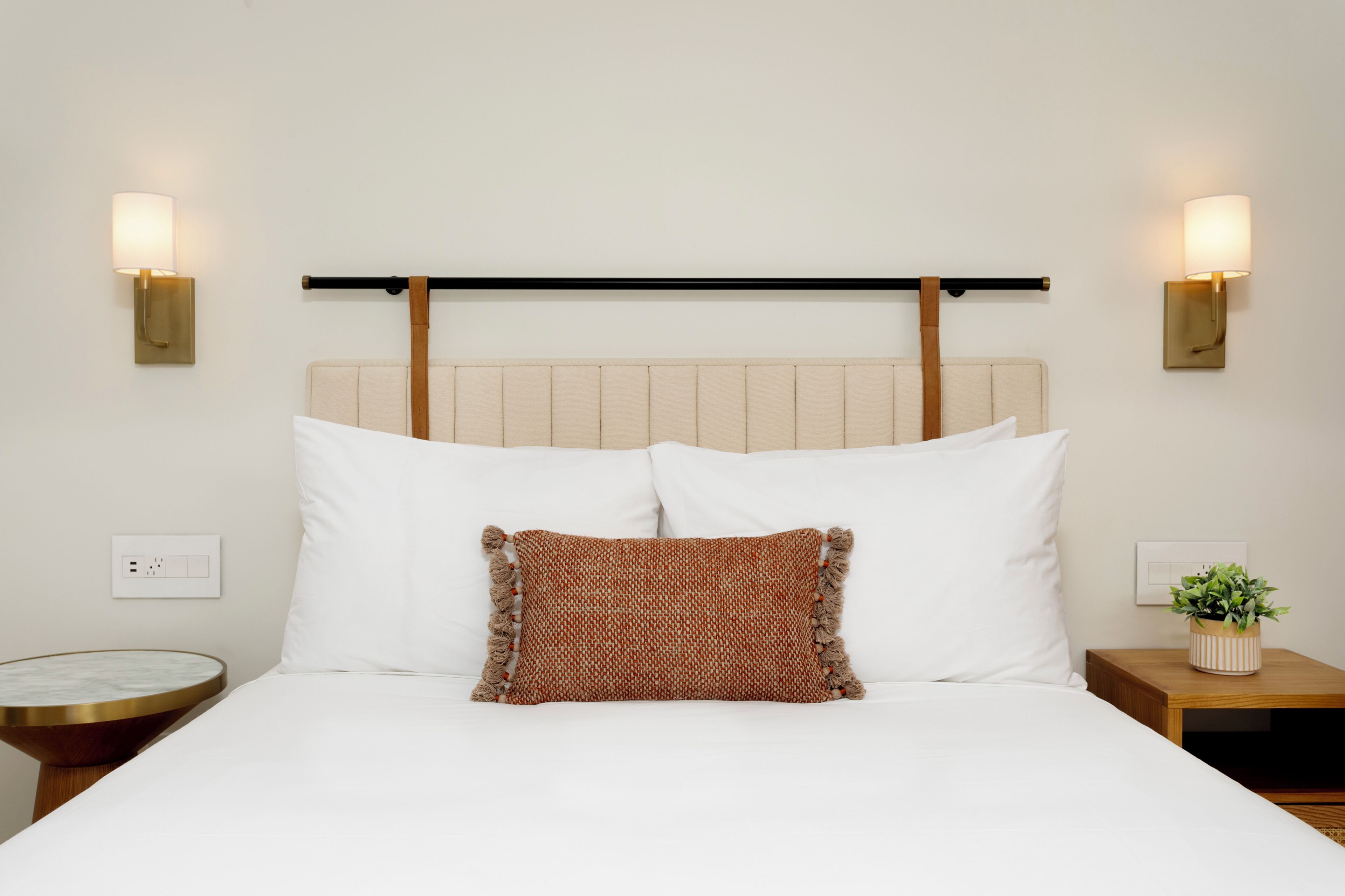 A hotel room with a bed, two wall-mounted lamps, a headboard, a bedside table, and a nightstand with a plant.