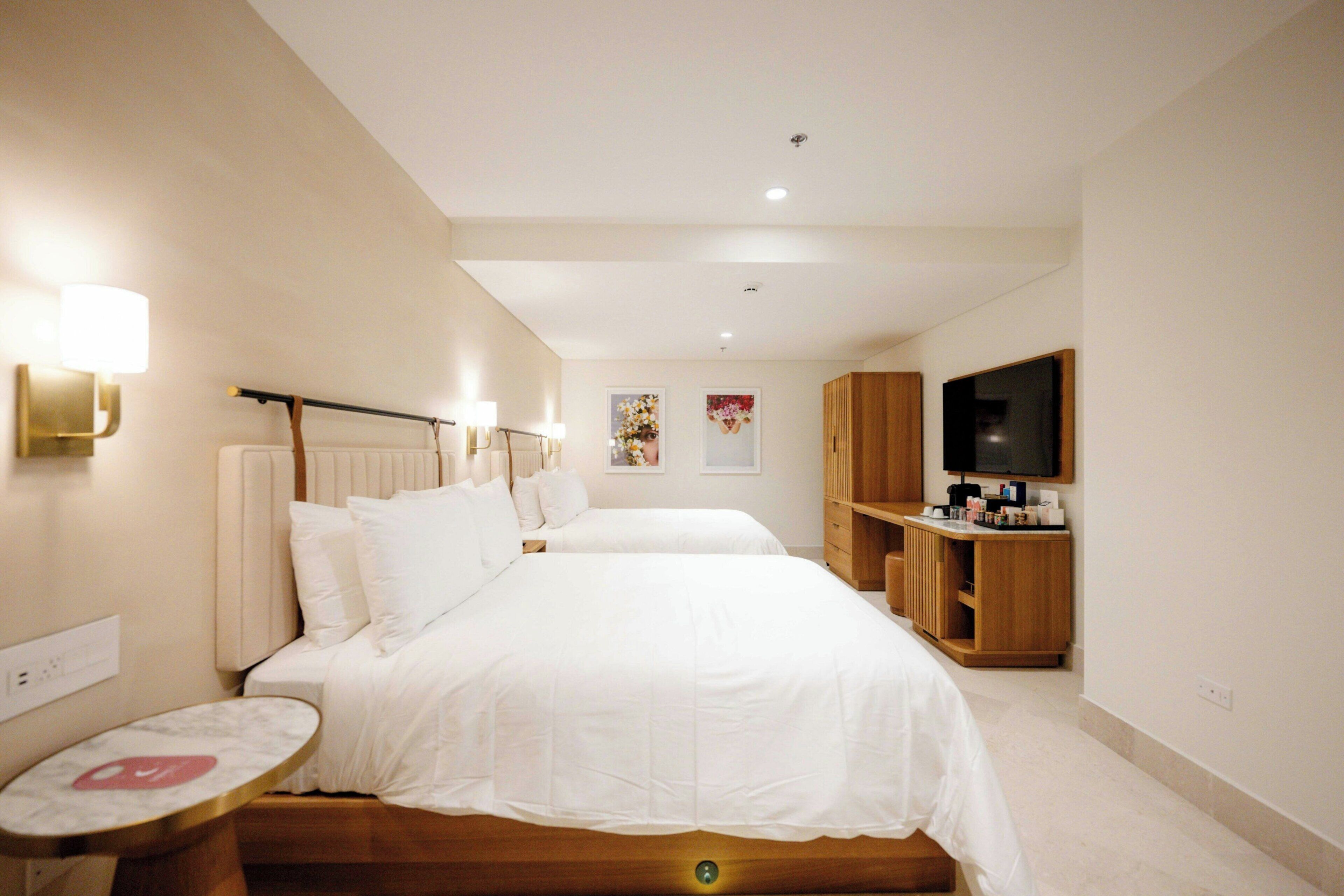 Quarto de hotel com duas camas, uma televisão, um armário de madeira e uma mesinha.