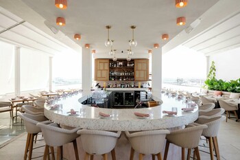 A modern hotel room with a large bed, a seating area with a chair and a small table, and a view of the sea through arched windows. at Alma San Juan, Puerto Rico, Autograph Collection