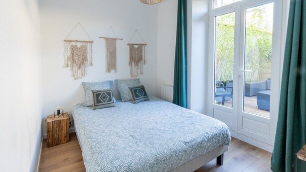 A bedroom with a bed, a wooden nightstand, macramé wall hangings, a hanging light fixture, and a view of an outdoor patio through a glass door.