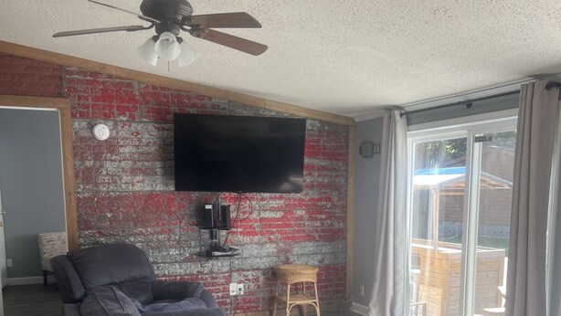 Un salon avec un canapé noir, une table basse en bois, un ventilateur de plafond, un téléviseur fixé au mur en briques et une porte-fenêtre coulissante donnant sur un espace extérieur.