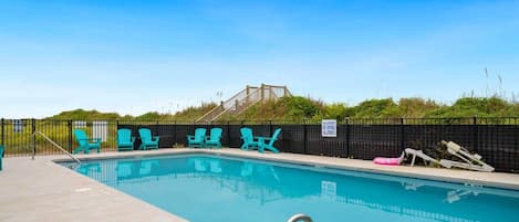Une piscine entourée d’une clôture noire, avec des chaises turquoise et un panneau indiquant qu’il est interdit de plonger.