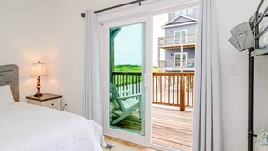 A bedroom with a bed, a nightstand with a lamp, a sliding glass door leading to a balcony with a view of a house and greenery.