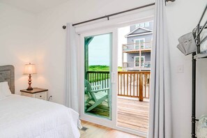 A bedroom with a bed, a nightstand with a lamp, a sliding glass door leading to a balcony with a view of a house and greenery.
