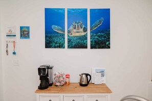 A wall with three framed pictures of sea turtles, a coffee maker, a kettle, a jar of jam, and a whiteboard with writing.