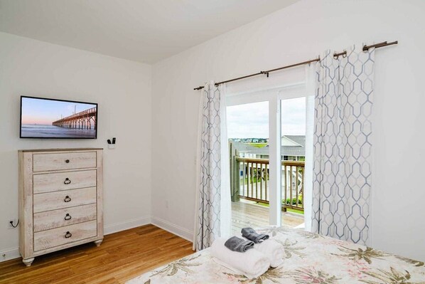 A bedroom with a wooden dresser, a bed with floral bedding, a television on the wall, and a view through a glass door to an outdoor deck.