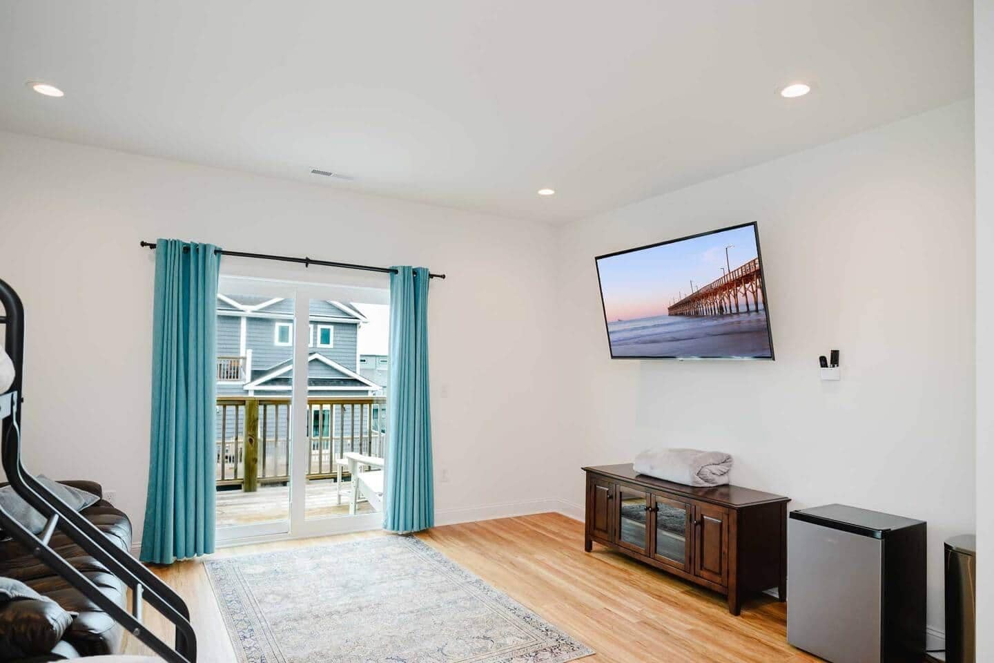 A room with a large sliding glass door leading to a balcony, a flat-screen TV mounted on the wall, a wooden cabinet, and a small refrigerator.