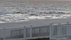 Escena costera con un atardecer sobre el océano, un muelle de madera y una valla blanca.