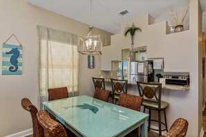 Un comedor con una mesa de vidrio y sillas de mimbre, una lámpara colgante y una cocina al fondo.