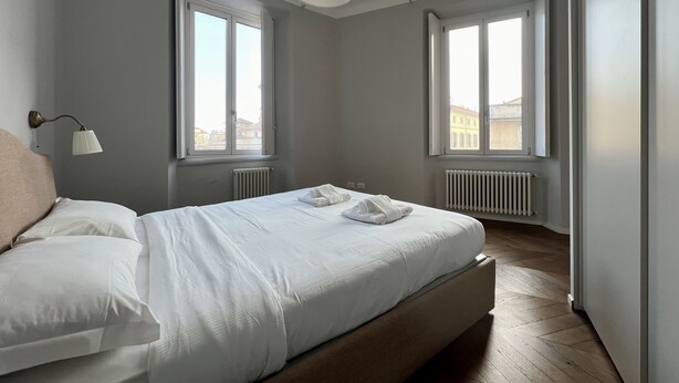 Un dormitorio con cama, dos ventanas, un radiador y una lámpara colgante.