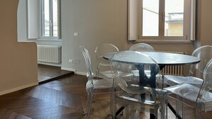 Un comedor con una mesa redonda y sillas transparentes, ventanas amplias y un radiador.