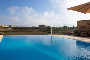 An infinity pool with a clear view of the countryside, a lounge area with chairs, and a large umbrella providing shade. - Kenn Holiday Home 4 bedrooms with private pool (L-Għarb)