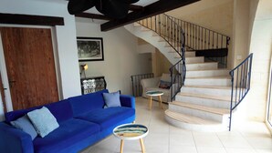 Un salon avec un canapé bleu, deux petites tables rondes, un escalier avec une rampe noire et une porte en bois.