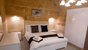 A bedroom with a white bed, two nightstands, a wardrobe, and a stone wall. - SurWig Vacation Townhouse (Kerċem)