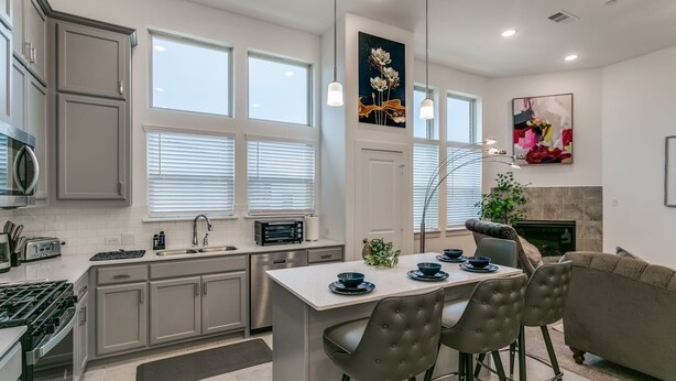 A modern kitchen with grey cabinets, a central island, and a dining area with a grey sofa and bar stools. - Home away from home  (Lewisville)