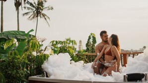 Een koppel in een jacuzzi, omringd door schuim, met palmbomen en een gebouw op de achtergrond.