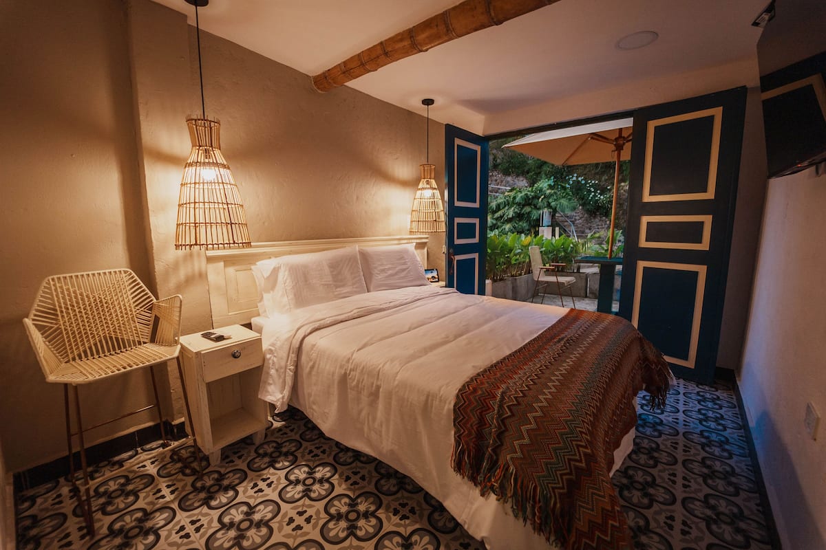 Ein Schlafzimmer mit Bett, Nachttisch, Stuhl, einem gemusterten Boden und Blick ins Freie.
