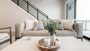 A modern living room with a beige sofa, a coffee table with a candle and decorative items, a staircase with black railings, and a large window.