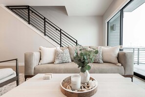 A modern living room with a beige sofa, a coffee table with a candle and decorative items, a staircase with black railings, and a large window. - Hideout Epic View • Modern Home • 10 Min to Deer Valley • Lake & Mountain Views (Hideout)