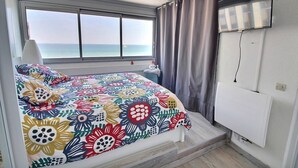 Ein Schlafzimmer mit Bett, Fernseher, einem Fenster mit Vorhängen und Meerblick.