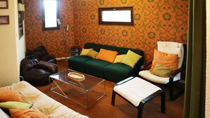 Un salon avec un canapé vert, un fauteuil blanc, une table basse en verre et un pouf en cuir marron.