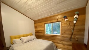 A bedroom with a bed, wooden walls, a window, and a wooden ceiling. - Beachside Cottage - on Lake Superior  (Cornucopia)