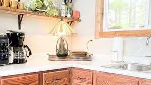 A kitchen with wooden cabinets, a coffee maker, a lamp, and a window with a view of trees. - Beachside Cottage - on Lake Superior  (Cornucopia)