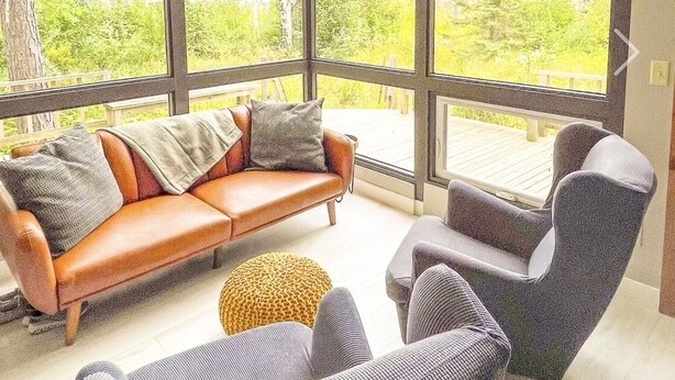 A modern living room with a large glass wall, a brown leather sofa, two armchairs, and a small round ottoman. - Beachside Cottage - on Lake Superior  (Cornucopia)