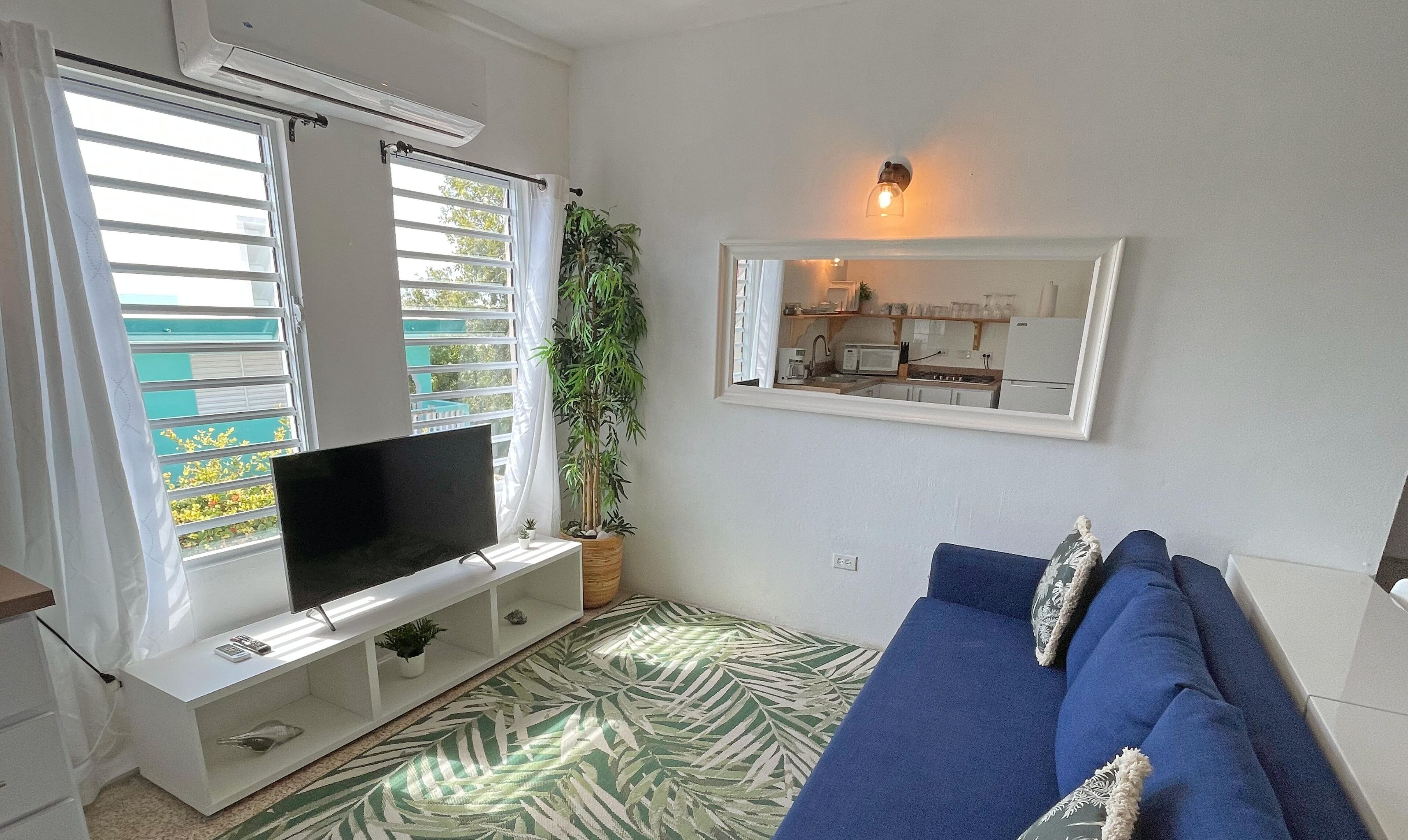 Ein Wohnzimmer mit Fernseher auf einem weißen Schrank, einem blauen Sofa, einem gemusterten Teppich, einer Topfpflanze und einer durch ein Fenster sichtbaren Küche.