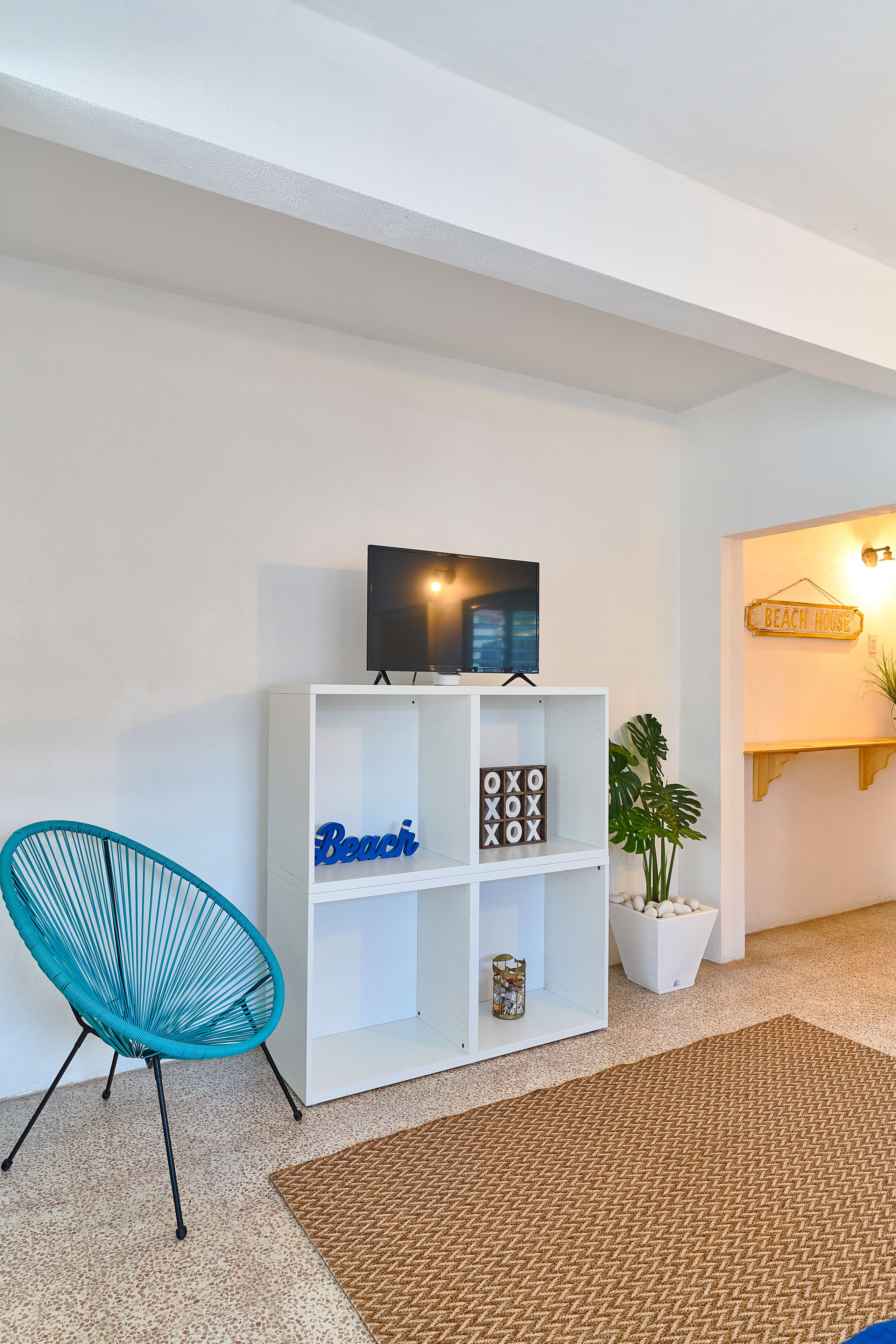 Una sala moderna con una estantería blanca, un sillón azul, un televisor de pantalla plana y un letrero en la pared que dice 'Beach House'.