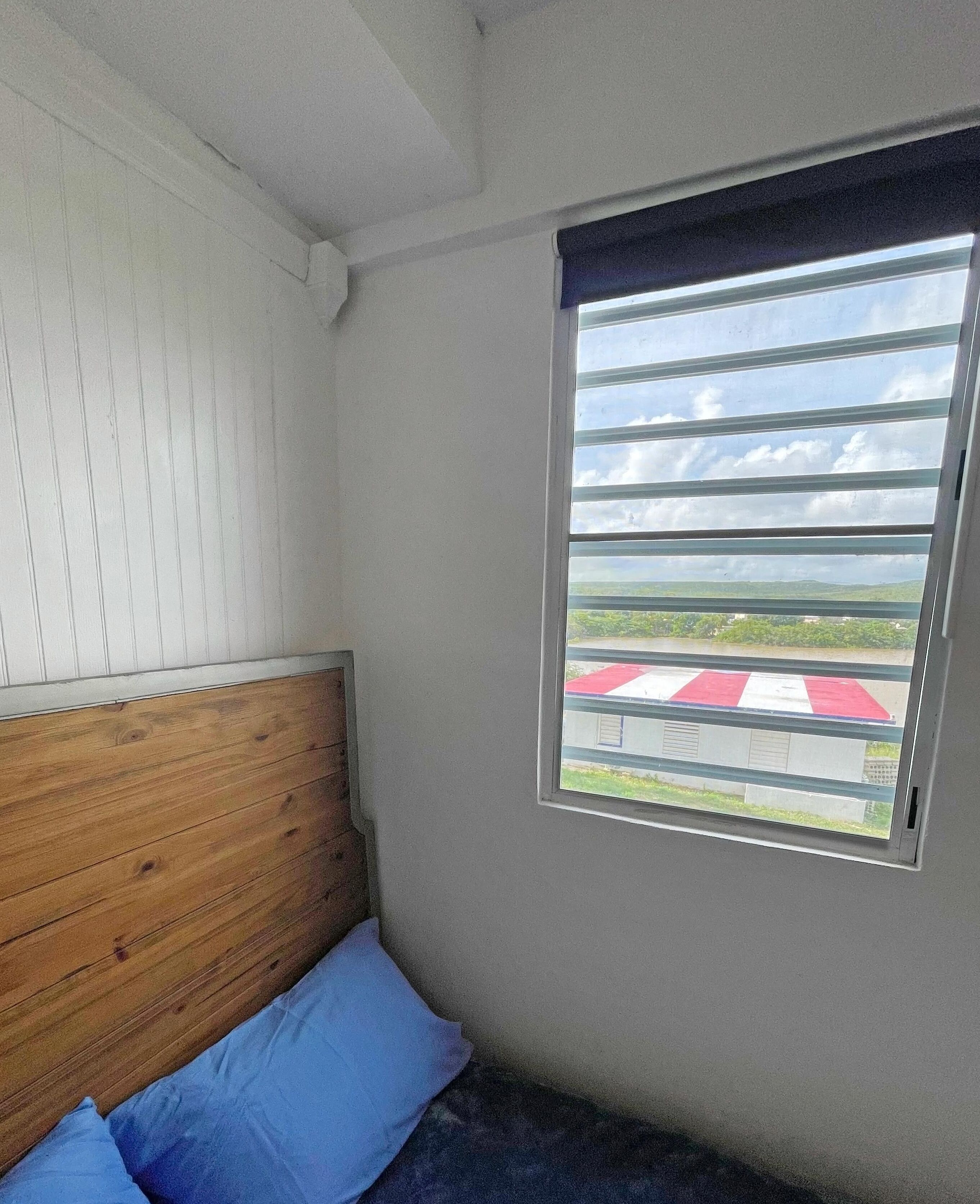 A room with a wooden headboard, a single bed with blue bedding, and a window with a view of a red and white checkered area outside.