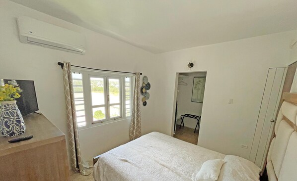 Une chambre à coucher avec un lit, une fenêtre avec des rideaux, un climatiseur fixé au mur, un miroir et une porte.