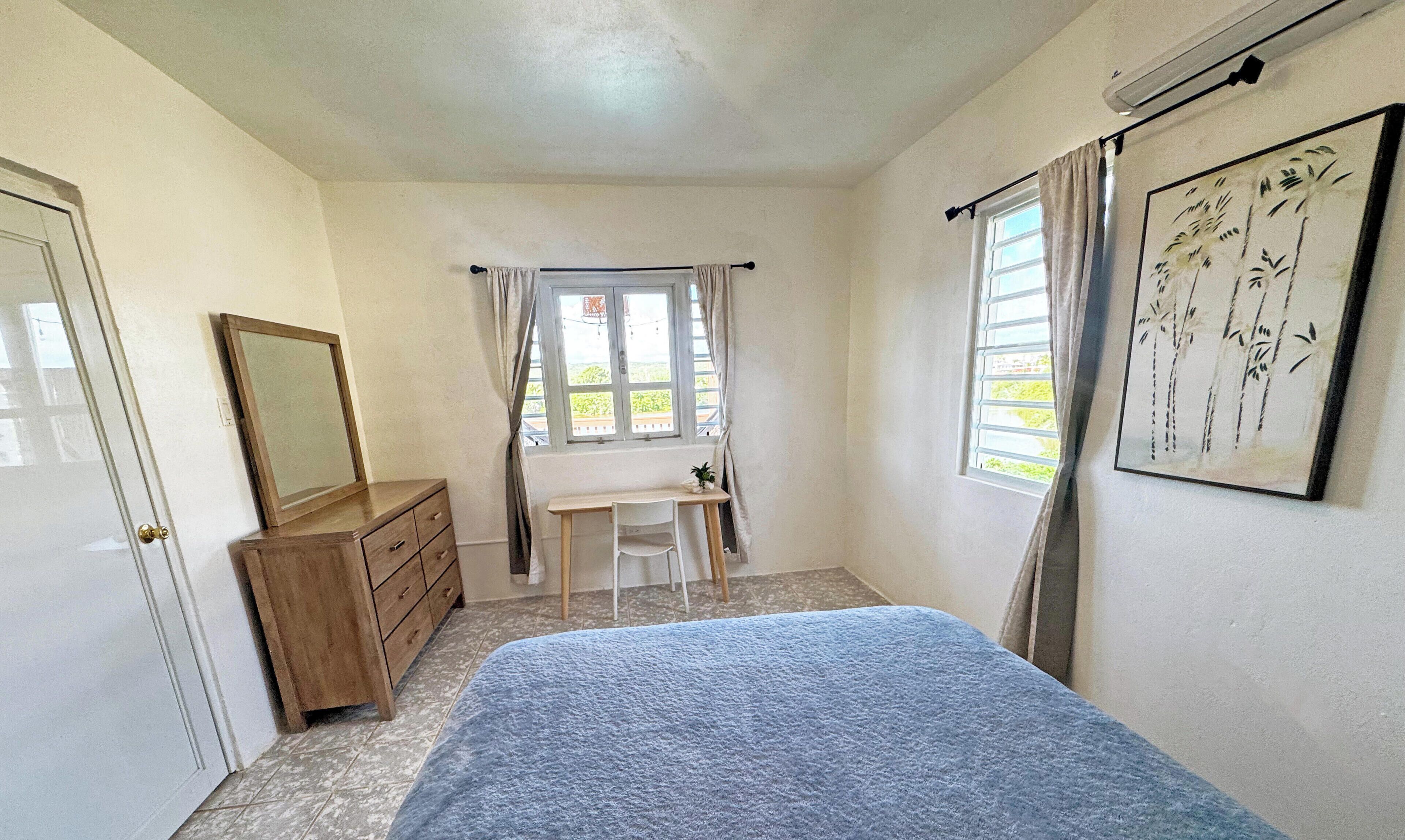 A bedroom with a bed, a dresser, a desk, two windows with curtains, and a wall-mounted artwork.