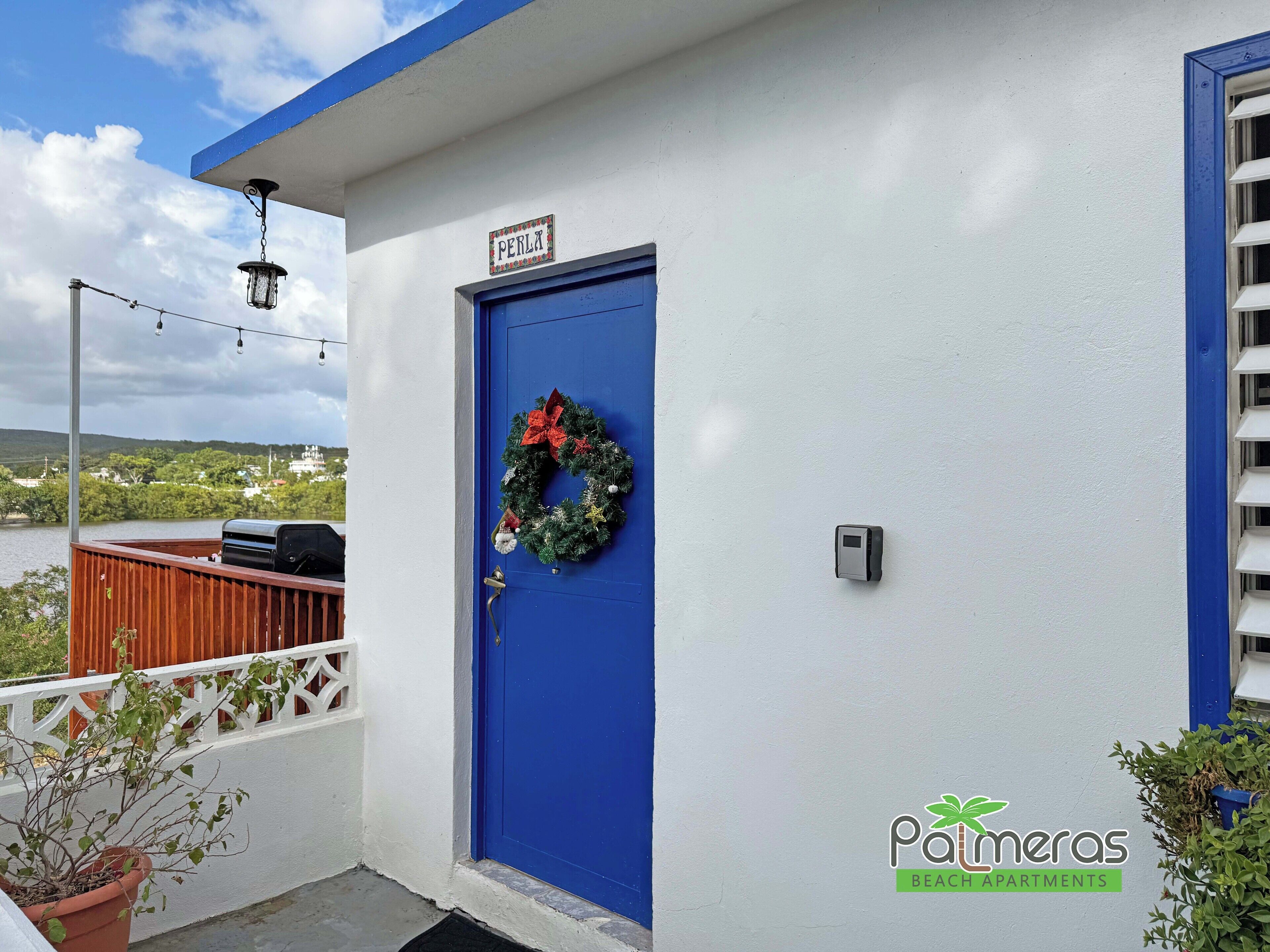 Una puerta azul con un adorno de laurel, un letrero que dice "PERLA" y el logotipo de "Palmeras Beach Apartments".