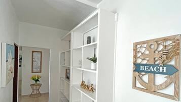 Um corredor com estantes brancas, uma pequena mesa com um vaso de flores e um enfeite de parede com a palavra 'BEACH'.