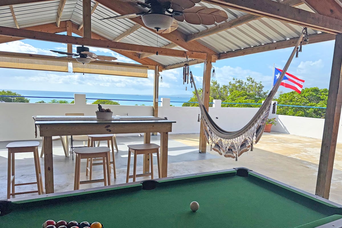 Une table de billard, un hamac suspendu et un drapeau dans un espace extérieur abrité, avec vue sur l’océan.