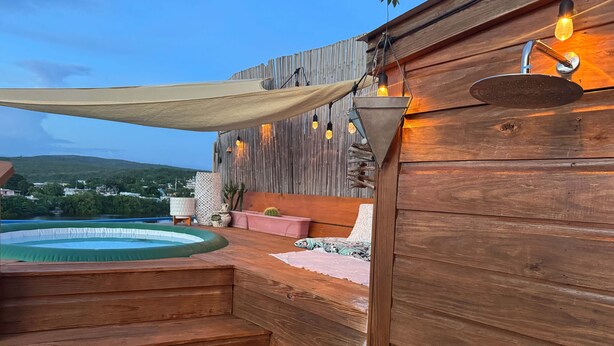 A wooden deck with a hot tub, a hammock, and a view of a town and hills. - Palmeras Beach Apartments (Guanica)