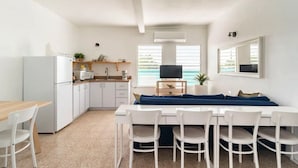 A modern kitchen with white cabinets, a dining table with chairs, a blue sofa, and a television on a wooden stand. - Palmeras Beach Apartments (Guanica)