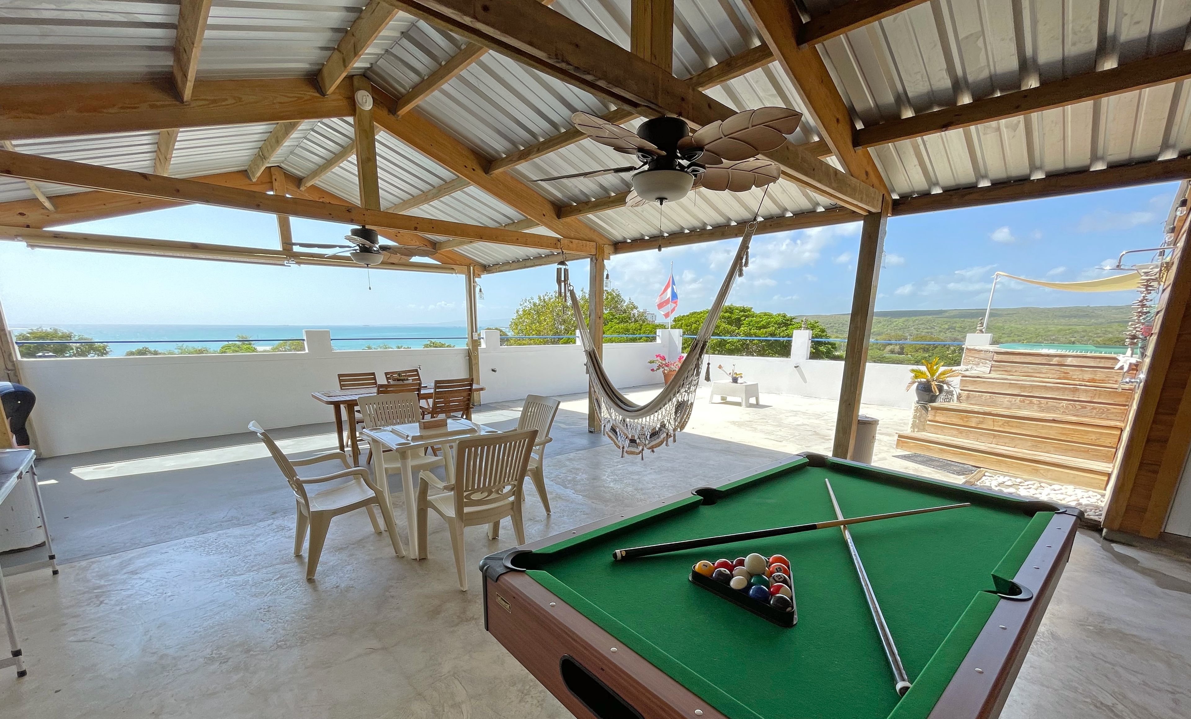 A pool table with pool balls, a hammock, and a table with chairs in a covered outdoor area with a view of the sea.