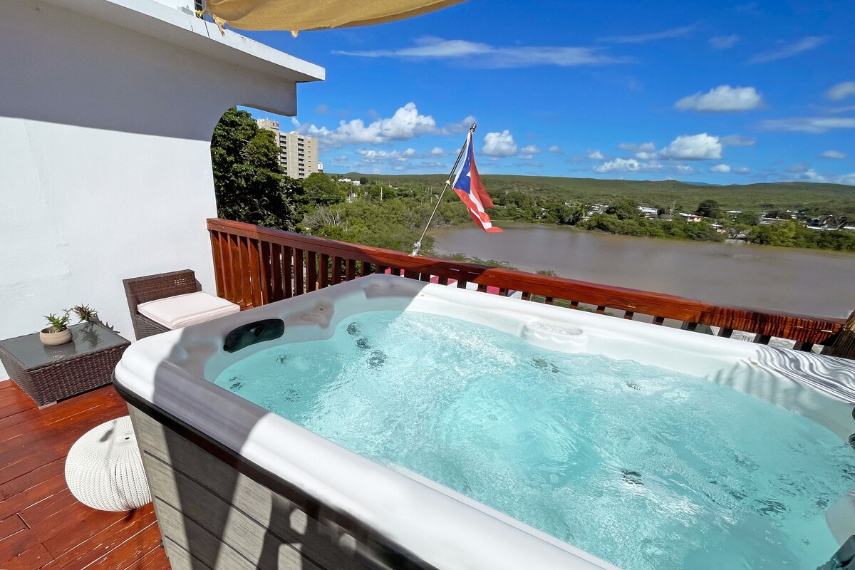 A hot tub on a wooden deck with a view of a river and hills.