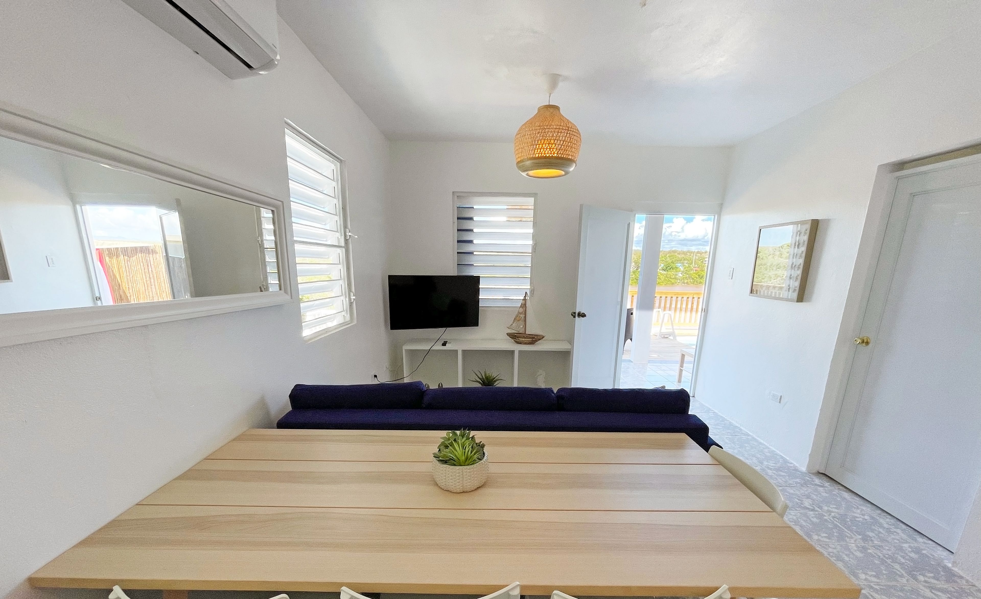 A modern living room with a wooden dining table, white chairs, a blue sofa, and a TV on the wall.