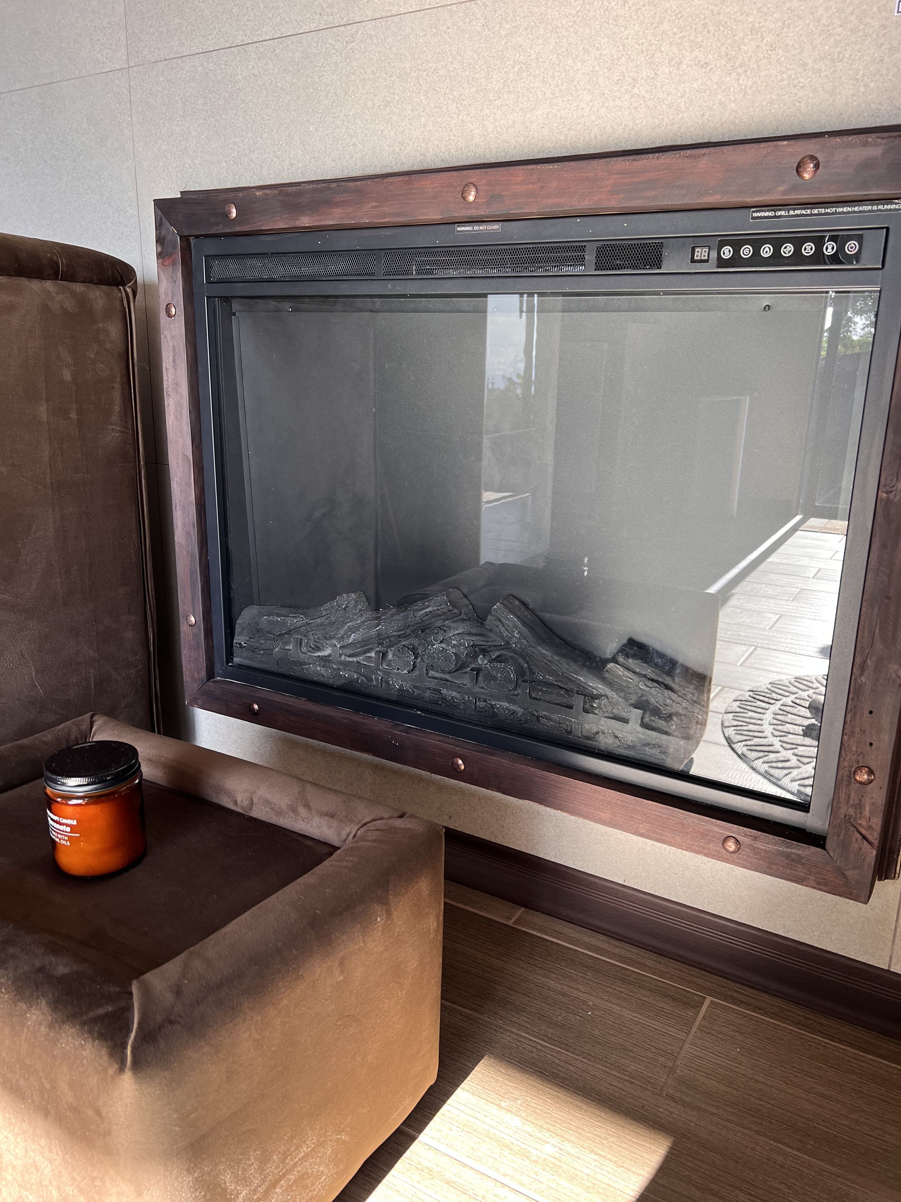A glass-enclosed fireplace with a wooden frame, a brown cushioned bench, and a candle on the bench.