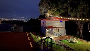 A small, single-story house with a red roof, a visible window, and a porch area with outdoor furniture. - Villazul 2 (Sabana Redonda)
