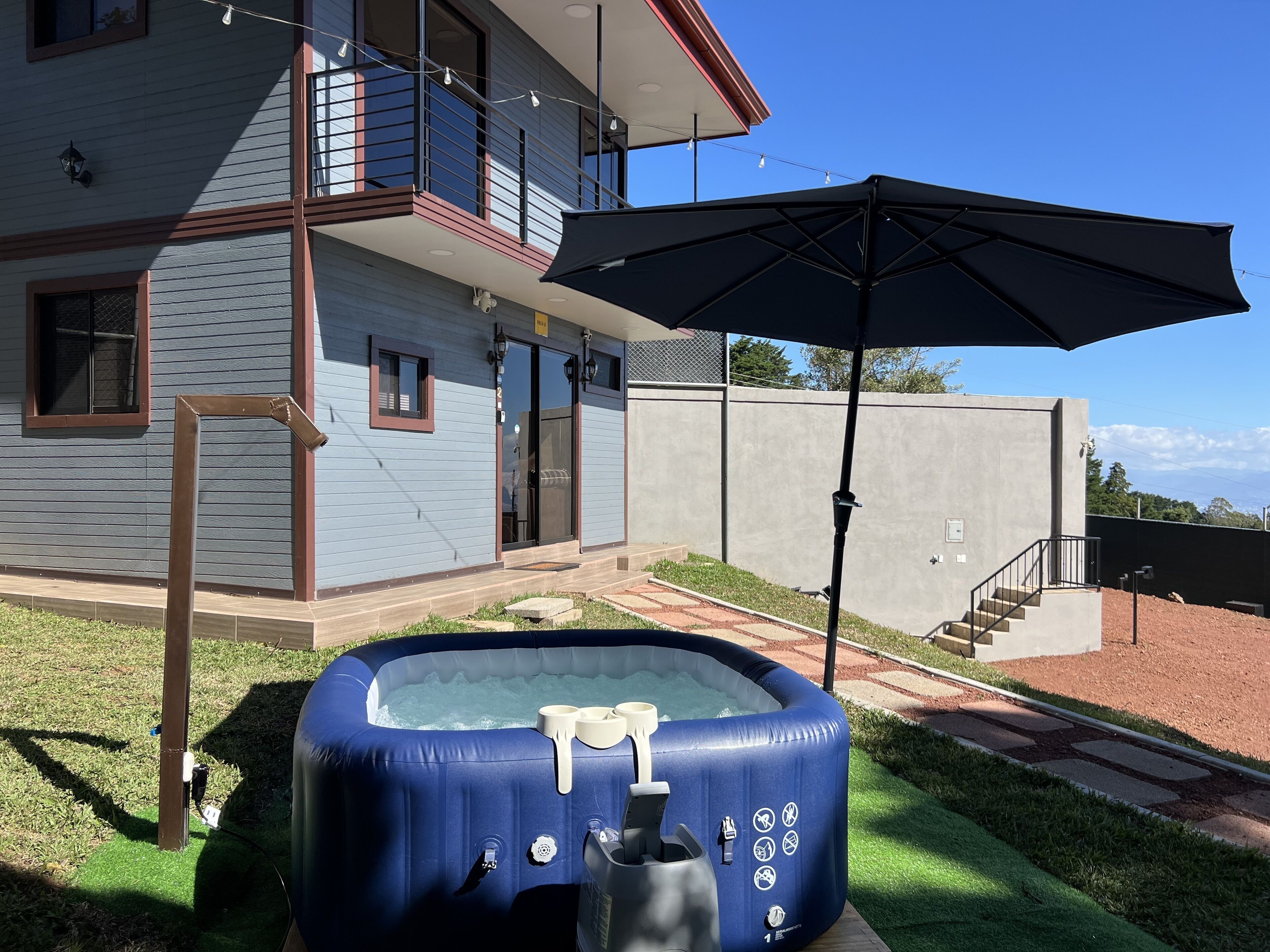 Un jacuzzi gonflable bleu, avec une pompe et des gobelets, un parasol de patio et une maison dotée d’un balcon et d’une porte.