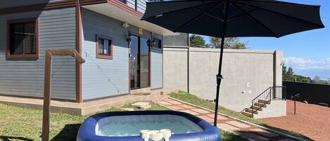 Un jacuzzi gonflable bleu, avec une pompe et des gobelets, un parasol de patio et une maison dotée d’un balcon et d’une porte.