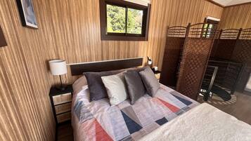 Una cama bien hecha con un edredón estampado, una lámpara de noche y una ventana con vistas a la vegetación.